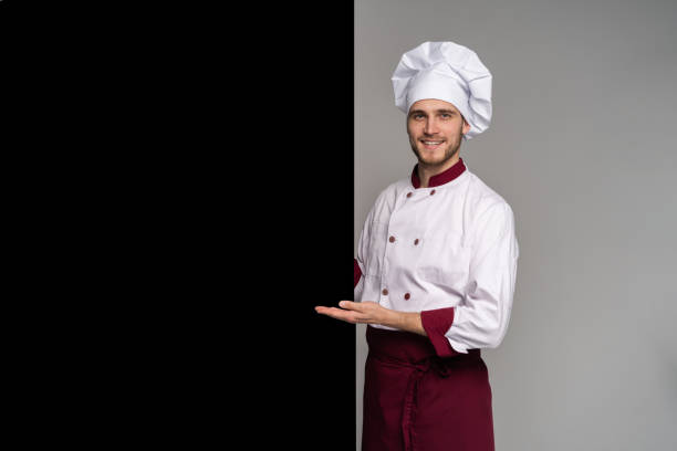 istock Image of smiling young cook in uniform standing isolated over grey wall background. Looking camera pointing. 1166608485