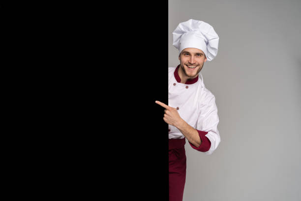 istock Image of smiling young cook in uniform standing isolated over grey wall background. Looking camera pointing. 1166608494
