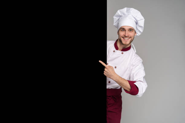 Image of smiling young cook in uniform standing isolated over grey wall background. Looking camera pointing.