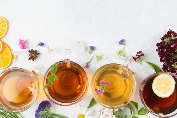 Directly above view of a four tea cups with different herbal teas. Anise, flowers and dried orange slices are spread around the tea cups on white background. One of the teas has a lemon slice.