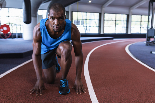 Portrait of African-american male athletic in starting position on running track in fitness center. Bright modern gym with fit healthy people working out and training