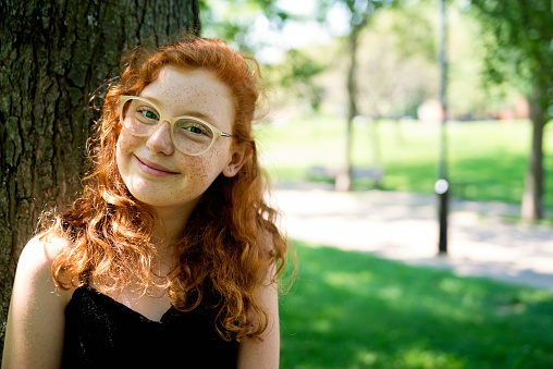 Portrait of beautiful teenage red hair girl in a park on a sunny day. She has curly hair, freckles, eyeglasses and is looking at the camera with a smile. Horizontal waist up outdoors shot with copy space.