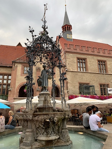 Göttingen, Germany - August, 1 - 2019:  The Gänseliesel Fountain in front of the Old Town Hall in downtown Göttingen.  The fountain with a goose-girl as a fountain figure was built in 1901 and is considered a landmark of the university city. The bronze figure was created by the sculptor Paul Nisse after a design by the architect Heinrich Stöckhardt. Since 1990, the fountain figure is a copy, while the original figure is in the Municipal Museum. Some people sitting on the fountain, talking and having fun.