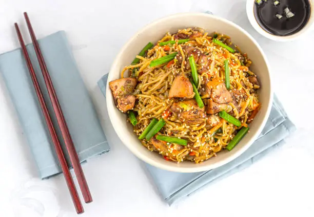 Stir-Fried Noodles with Chicken and Vegetables in a White Bowl with Chopsticks Directly Above Photo.