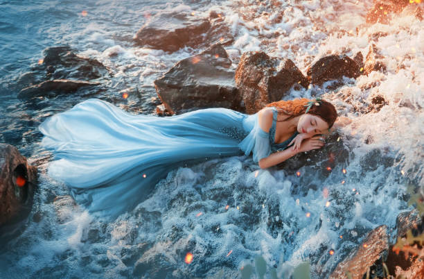 濡れた石の上に休む魅力的な海の王女、青い長い柔らかいドレスの女の子は甘く頭の下に手で滝の冷たい水の中で眠る、暖かい太陽の光の中で親切な顔を持つ甘い赤い人魚 - 人魚 ストックフォトと画像