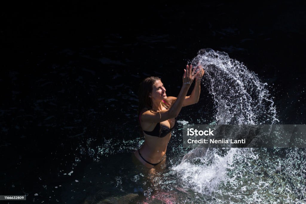 Woman splashing sea water Young woman in bikini having fun splashing water at sea at rocky beach in shade Adult Stock Photo