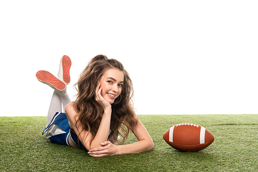 happy cheerleader girl in blue uniform lying near rugby ball on green field isolated on white