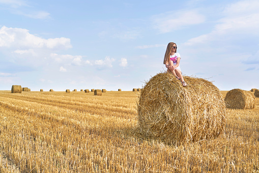 photograph of a blonde girl in sunglasses on a circular straw bale in a wheat field in the sun in Belarus