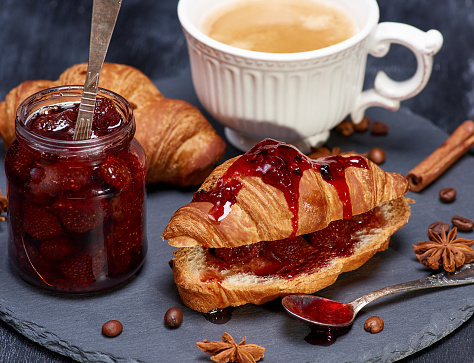 croissant with strawberry jam and white cup with coffee on black surface