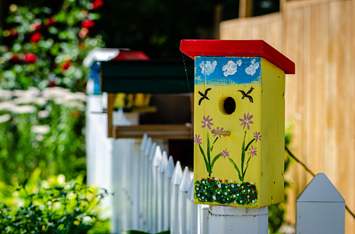 Close-up of yellow painted birdhouse on a picket fence with out of focus flowers in the background