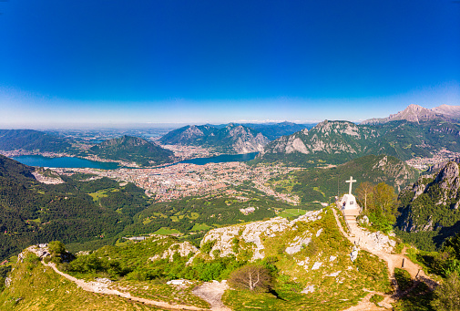 Beautiful panoramic sunrise landscape at town Lecco, lake Como in sunny summer day. Stunning airial view to cross on top of mountain from drone near viewpoint Piani Pizzo d'Erna, alps, italy, Europe