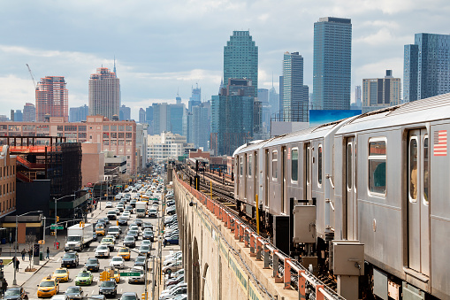 Train approaching  elevated subway station in Queens, New York. Financial buildings and New York skyline are seen in the background, on the left below can be seen a busy street full of cars at rush hour, USA.