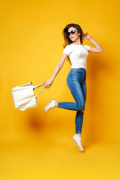 Beautiful Young Woman In Sunglasses White Shirt Blue Jeans Jumping With Bag  On The Yellow Background Stock Photo - Download Image Now - iStock