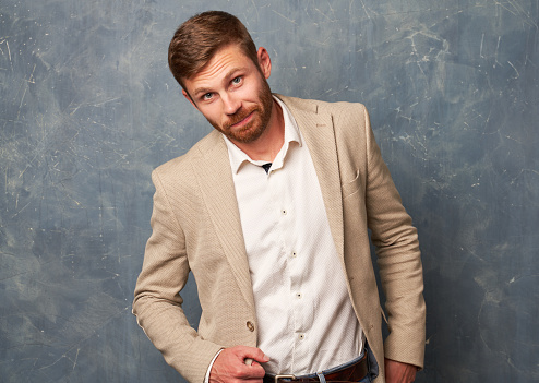 Young businessman wears elegant shirt with jacket. Male with hairstyle and ginger beard being in good mood isolated on blue textured background. Confidently from under forehead looking at camera.