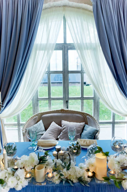 istock Wedding table decorated with white flowers, candles. Soft sofa with cushions, large window.Blue and white 1166675968