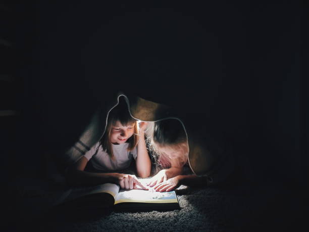 two sisters are reading a book under the covers at night with a flashlight stock photo