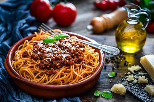 Clay plate filled with spaghetti with Bolognese sauce shot on rustic wooden table. Some ingredients like ripe tomatoes, olive oil, basil, peppercorns and Parmesan cheese are all around the plate. XXXL 42Mp studio photo taken with SONY A7rII and Sony FE 90mm f2.8 Macro G OSS lens