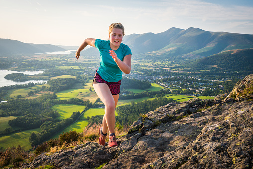 Female trail runner running up a rocky mountain crest overlooking the green valley below.