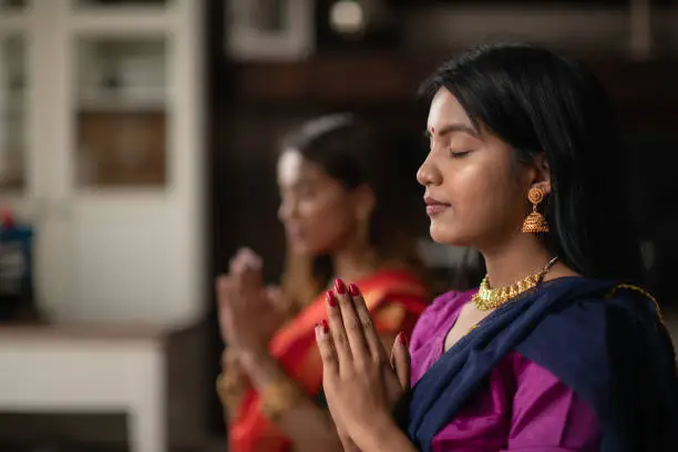 Two (2) Indian sisters are praying in their living room together. They are peaceful as they rest in prayer pose. They are connecting with their god and giving thanks during Diwali.