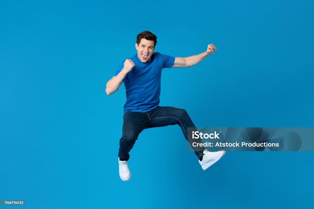 American man jumping and enyoying his success Smiling handsome American man joyfully jumping and raising his fists isolated on blue studio background  fro success and freedom concepts Men Stock Photo