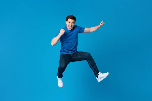 American man jumping and enyoying his success Smiling handsome American man joyfully jumping and raising his fists isolated on blue studio background  fro success and freedom concepts ecstatic photos stock pictures, royalty-free photos & images