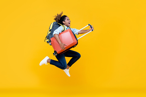 Excited African American woman tourist woman with backpack and luggage jumping in mid-air studio shot isolated on colorful yellow background