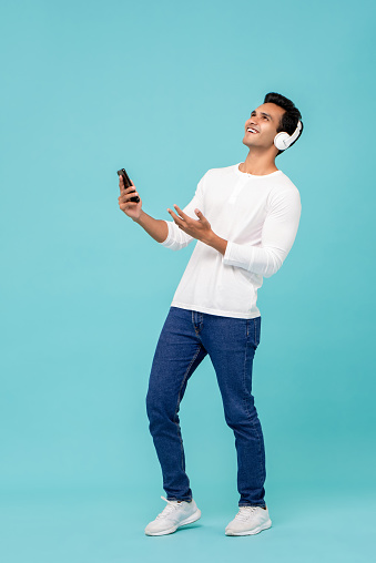 Smiling young Indian man in white t-shirt listening to music using wireless bluetooth headphones holding smartphone in hand isolated against blue background