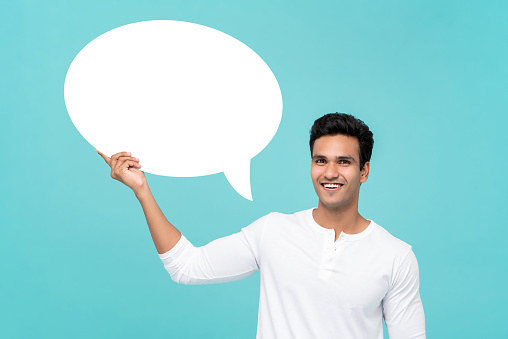 Handsome smiling Indian man in white t-shirt holding blank speech bubble sign isolated on light blue studio background