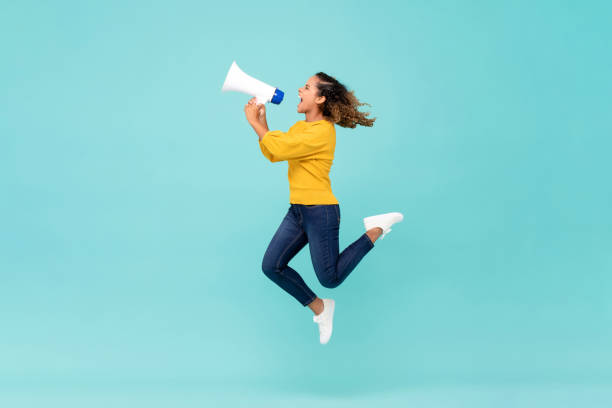 Girl with megaphone jumping and shouting Young African American girl with megaphone jumping and shouting on light blue background happy people audio stock pictures, royalty-free photos & images