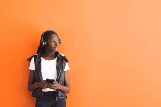 African Australian Teenage Girl Listening To Music Under Headphones Against Orange Wall In Urban Location