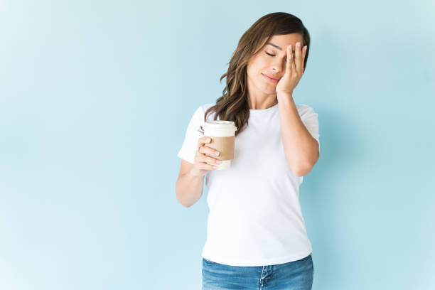 Tired Caucasian woman with coffee cup standing against blue background