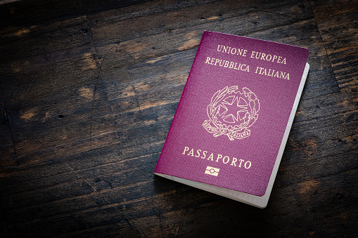 High angle view of Italian passport shot on dark wooden table. The composition is at the right of an horizontal frame leaving a useful copy space for text and/or logo at the left. Predominant colors are red and brown. Low key XXXL 42Mp studio photo taken with SONY A7rII and Sony FE 90mm f2.8 Macro G OSS lens