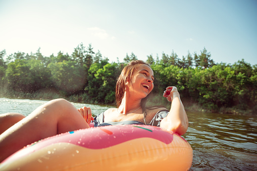 Happy woman having fun while laughting and swimming in river. Joyful female model with rubber ring as a donut at riverside in sunny day. Summertime, friendship, resort, weekend concept.