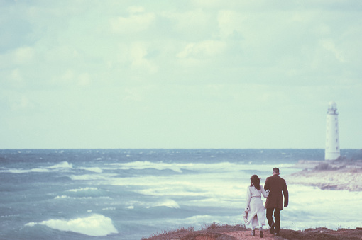 vintage picture of young couple in autumn coat walking along stormy sea shore with high waves and lonely lighthouse on edge of world