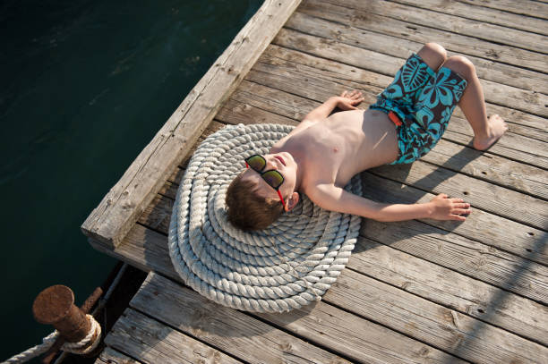 happiness concept of healthy idle little child in sunglasses and trendy shorts lying meditate on wood pier with coil of rope near sea water - fashionable the human body short hair human head imagens e fotografias de stock