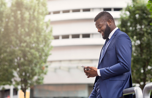 Attractive happy businessman is texting on cell phone near modern office building, side view with copy space. Business, technology and communication concept