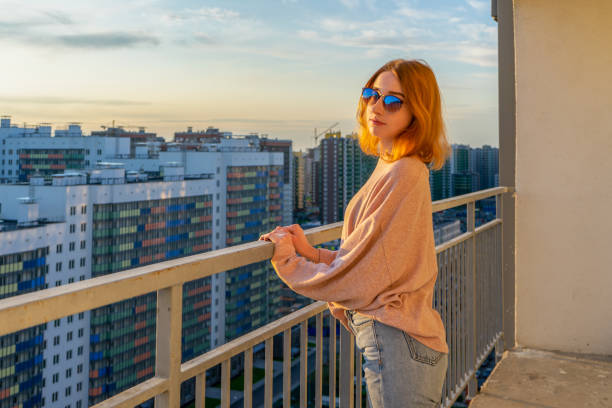 istock Tween redhead girl in pullover, jeans and sunglasses standing on balcony against high-rise multi-storey residential building at sunset. Beautiful look, fashionable city street outfit, teenage fashion 1166784013