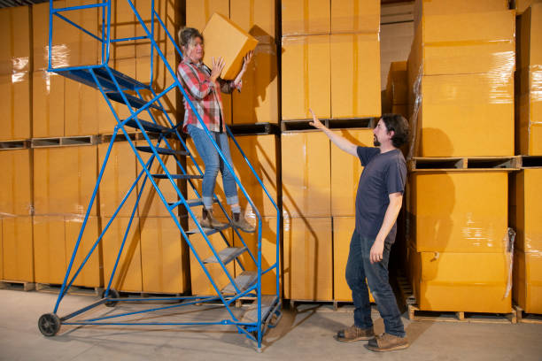 An industrial, warehouse, workplace safety topic. A female employee descending a rolling ladder unsafely.  She is not using three points of contact.  Her hand should be on the rail.  A coworker is warning her that she could risk a fall.