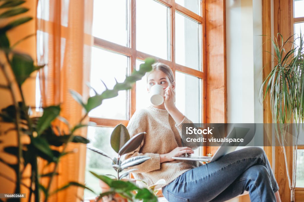 Candid portrait of woman drinking coffee Young woman drinking coffee, while working on her laptop. She's sitting by the window. Coffee - Drink Stock Photo