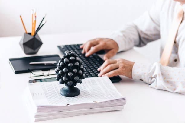 Close-up of hands middle-aged man in white shirt and yellow tie typing on keyboard laptop computer, concrete holder with pencils and pens, notebook, smartphone, glasses, stack of documents and black paperweight on white table, education office concept background primer plano de las manos de hombre de mediana edad con camisa blanca y corbata amarilla escribiendo en el ordenador portátil del teclado, soporte de hormigón con lápices y bolígrafos, cuaderno, teléfono inteligente, gafas y peso de papel negro en la