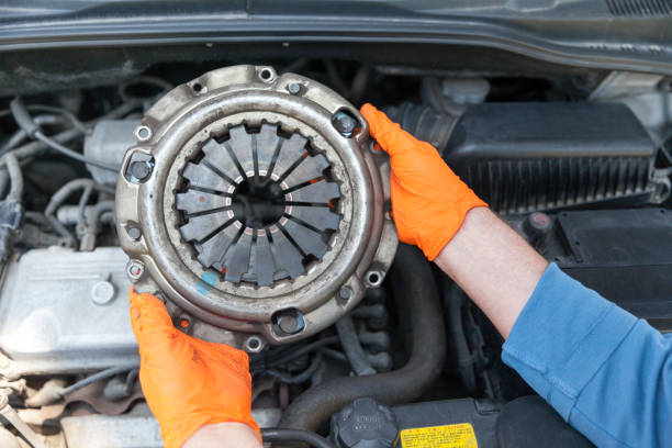 Auto mechanic wearing protective work gloves holding used clutch pressure plate above a car engine stock photo