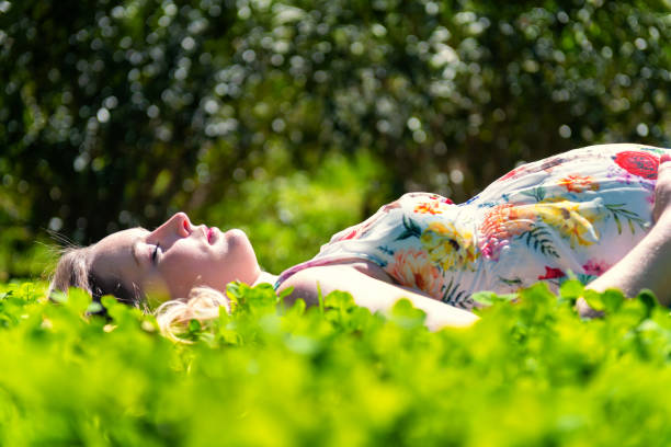 Beautiful pregnant woman lying down in grass and wearing floral dress. Young mother is surrounded by idyllic nature in summer. Future mother relaxing with eyes closed, napping, smiling and touching her belly. Beauty in life and lots of love for baby on the way.