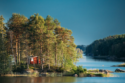 Sweden. Beautiful Red Swedish Wooden Log Cabin House On Rocky Island Coast In Summer Sunny Evening. Lake Or River Landscape.