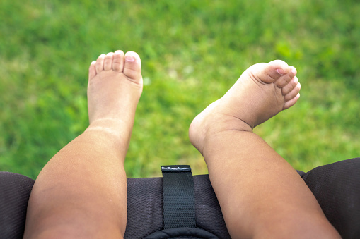 A close up of bare skin chubby legs, feet and toes of a mixed race baby boy infant hanging off into the fresh air over the grass at a park as he sits in his car seat stroller in the summer.