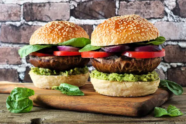 Portobello mushroom vegan burgers with avocado, tomato and spinach and onion on a wood serving board against a dark brick background