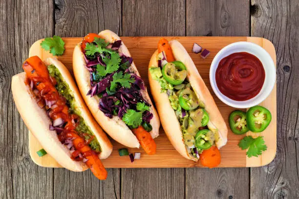 Carrot vegan hot dogs with assorted toppings. Top view on serving board with a rustic wood background. Plant based meatless meal concept.