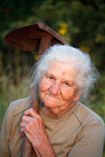 Closeup portrait of an old woman with gray hair smiling and looking at the camera, holding a rusty shovel in her hands, face in deep wrinkles, selective focus