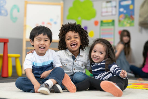 Happy Preschool Friends Portrait of three preschool age children. The multi-ethnic group of friends are embracing and laughing. The students are sitting together on the floor in their classroom. There are other school children playing in the background. summer camp photos stock pictures, royalty-free photos & images