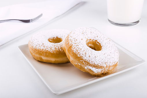 Donuts covered with icing sugar on a plate on a white background accompanied by a glass of milk and a fork on a napkin. Delicious breakfast or lunch. American food. Bakery and pastry products.