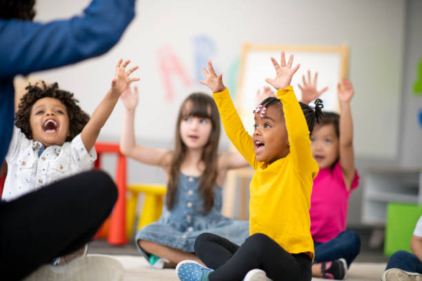 Multi-ethnic group of preschool students in class A multi-ethnic group of preschool students is sitting with their legs crossed on the floor in their classroom. The mixed-race female teacher is sitting on the floor facing the children. The happy kids are smiling and following the teacher's instructions. They have their arms raised in the air. child care stock pictures, royalty-free photos & images